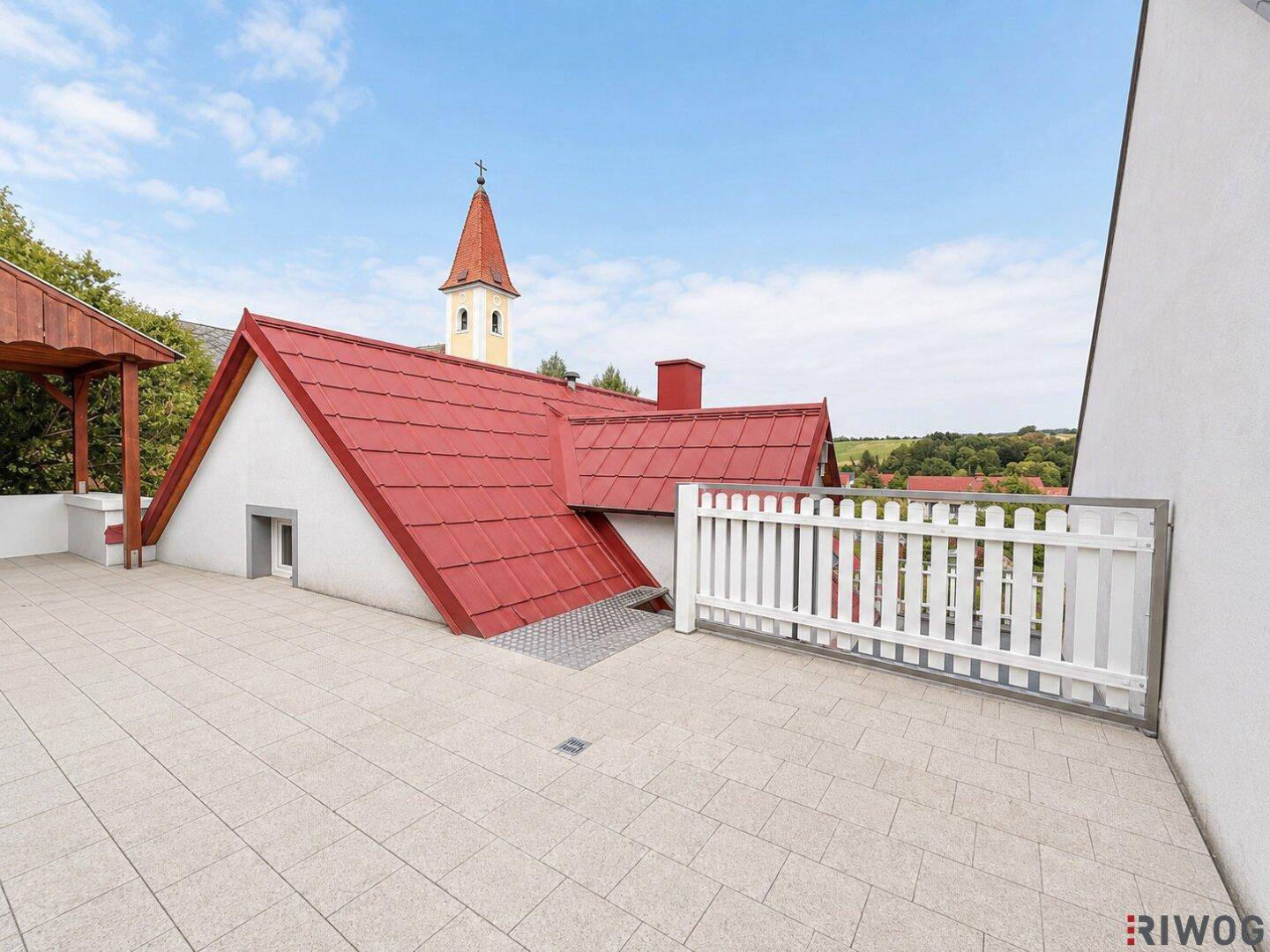 Geräumige Dachterrasse mit Fliesenboden und Blick auf die umliegende Landschaft und Kirche.