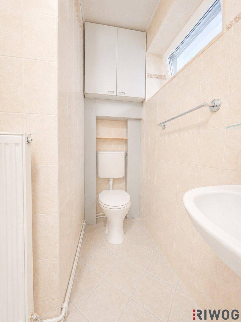 Kleines WC mit beigefarbenen Fliesen, Toilette, Waschbecken und einem schmalen Fenster.