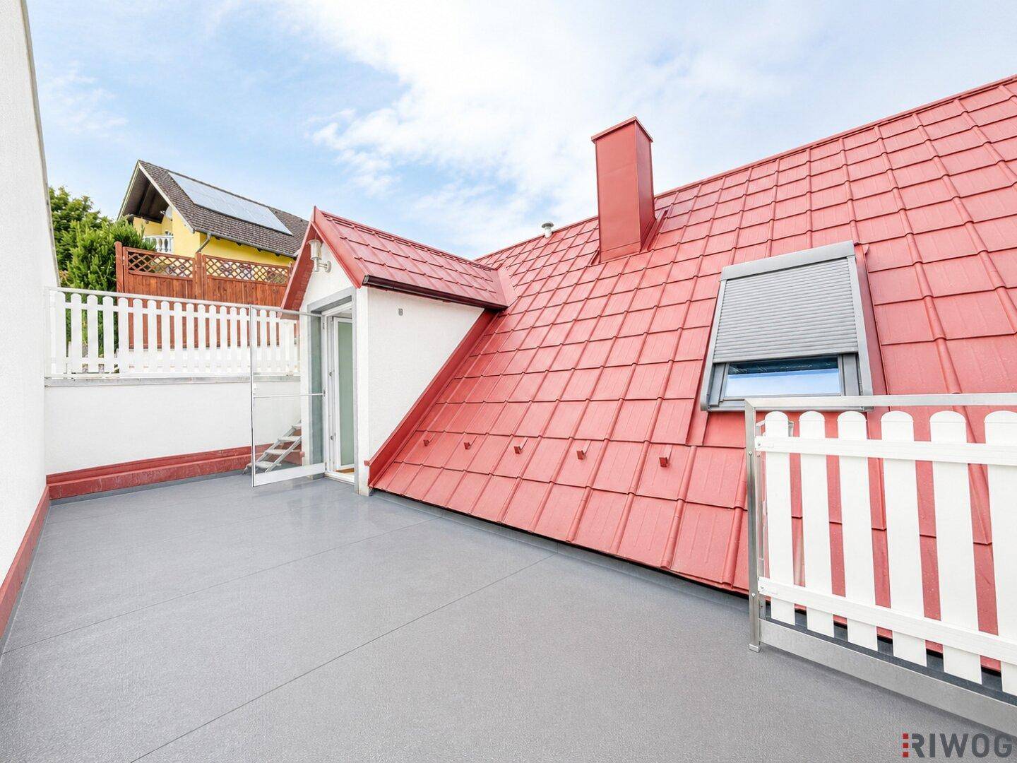 Geräumige Dachterrasse mit grauem Boden, weißem Geländer und Blick auf rote Ziegeldächer.