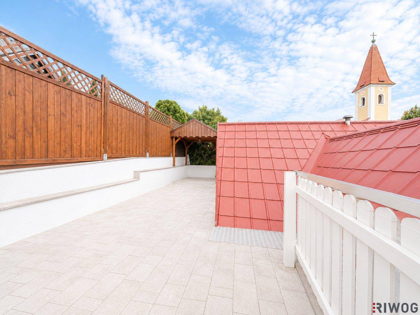 Weitläufige Dachterrasse mit Holzzaun, gepflastertem Boden und Blick auf einen Kirchturm.