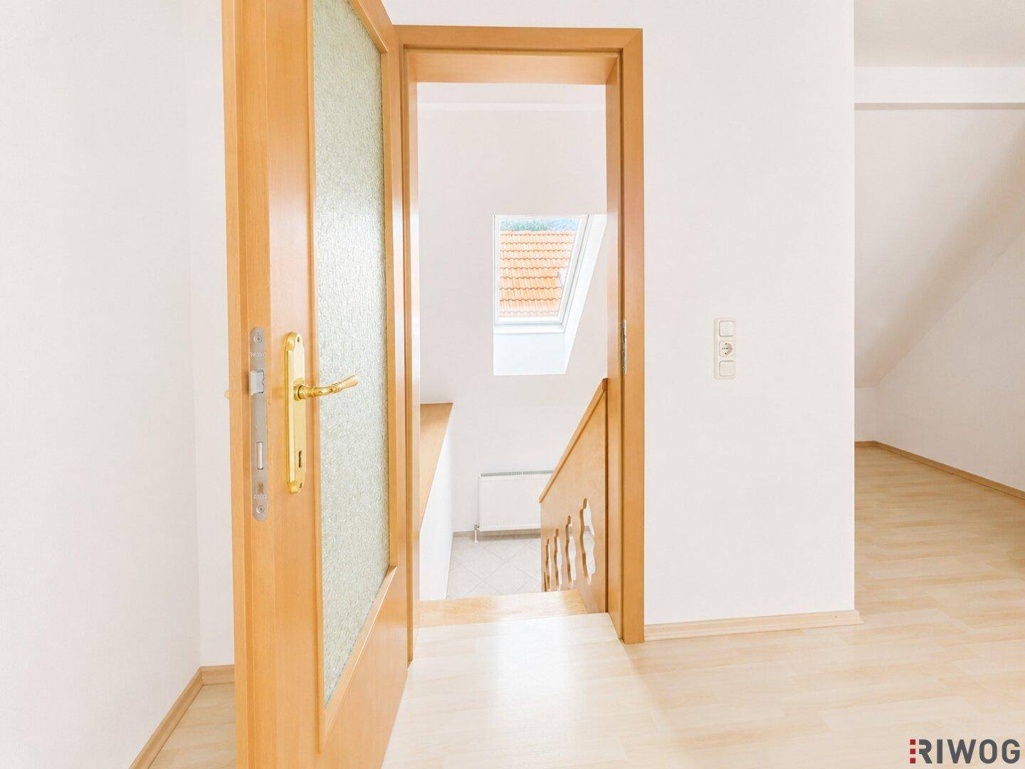 Offene Holztür führt zu einer Treppe mit Blick auf ein Dachfenster und den Himmel.