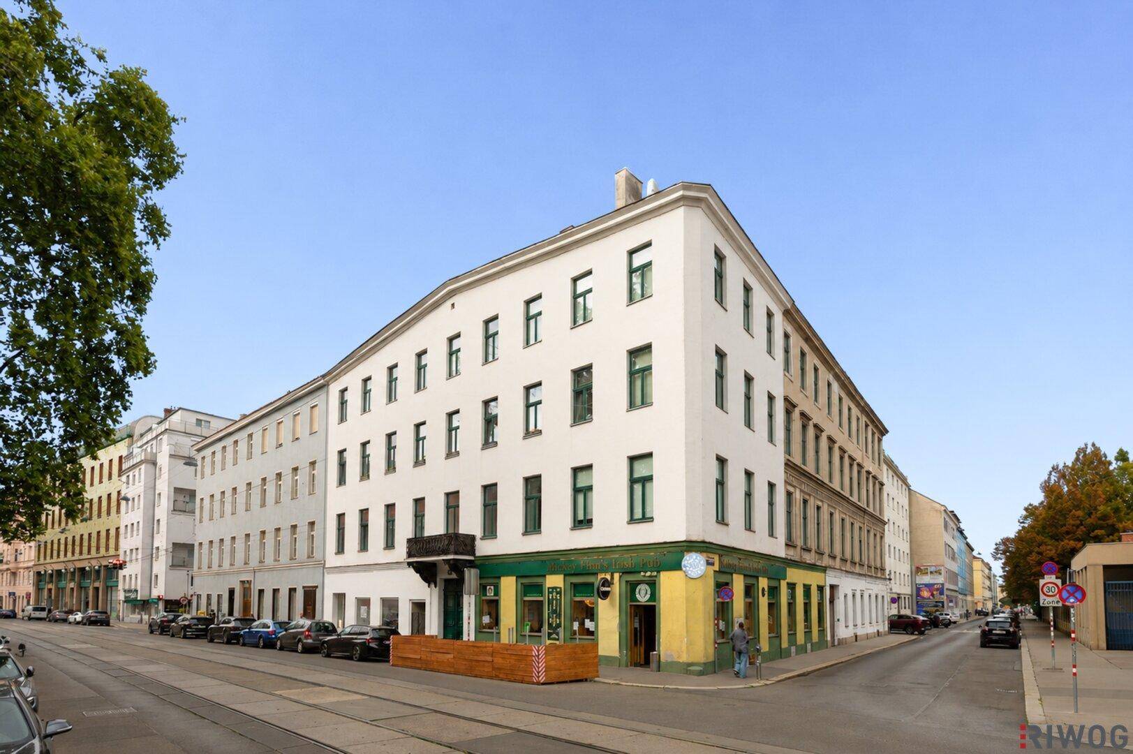 Eckgebäude mit klassischer Architektur, vielen Fenstern und Geschäften im Erdgeschoss an einer Straße.