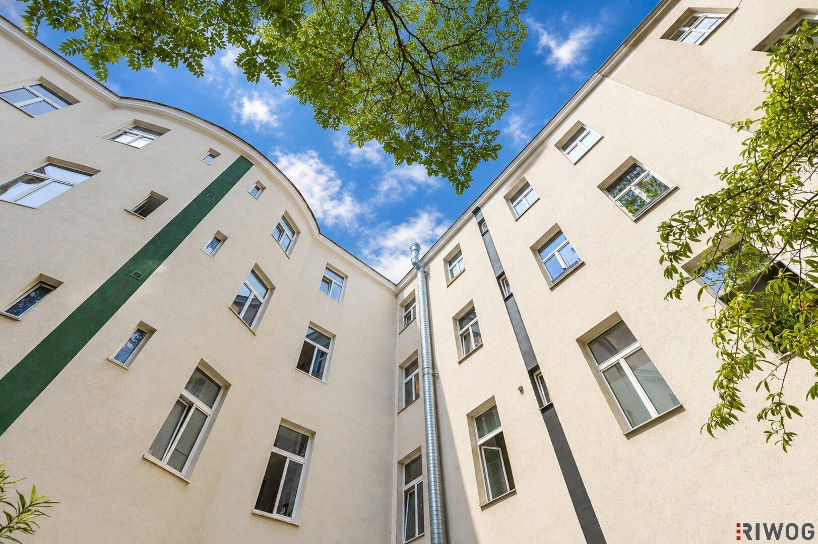 Blick nach oben in einen hellen Innenhof mit vielen Fenstern und grünen Bäumen.