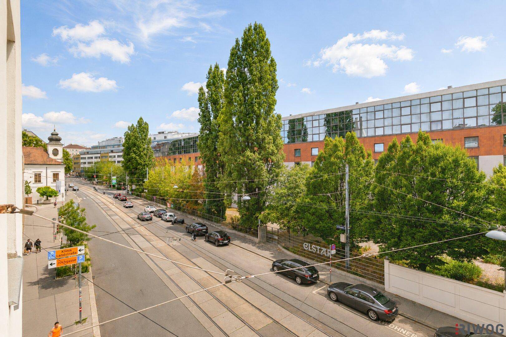 Weitläufiger Blick auf eine belebte Straße mit Bäumen und Gebäuden unter blauem Himmel.