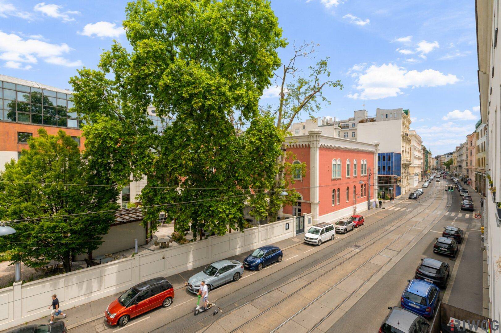 Panoramablick auf eine städtische Straße mit Gebäuden, Bäumen und vorbeifahrenden Autos.