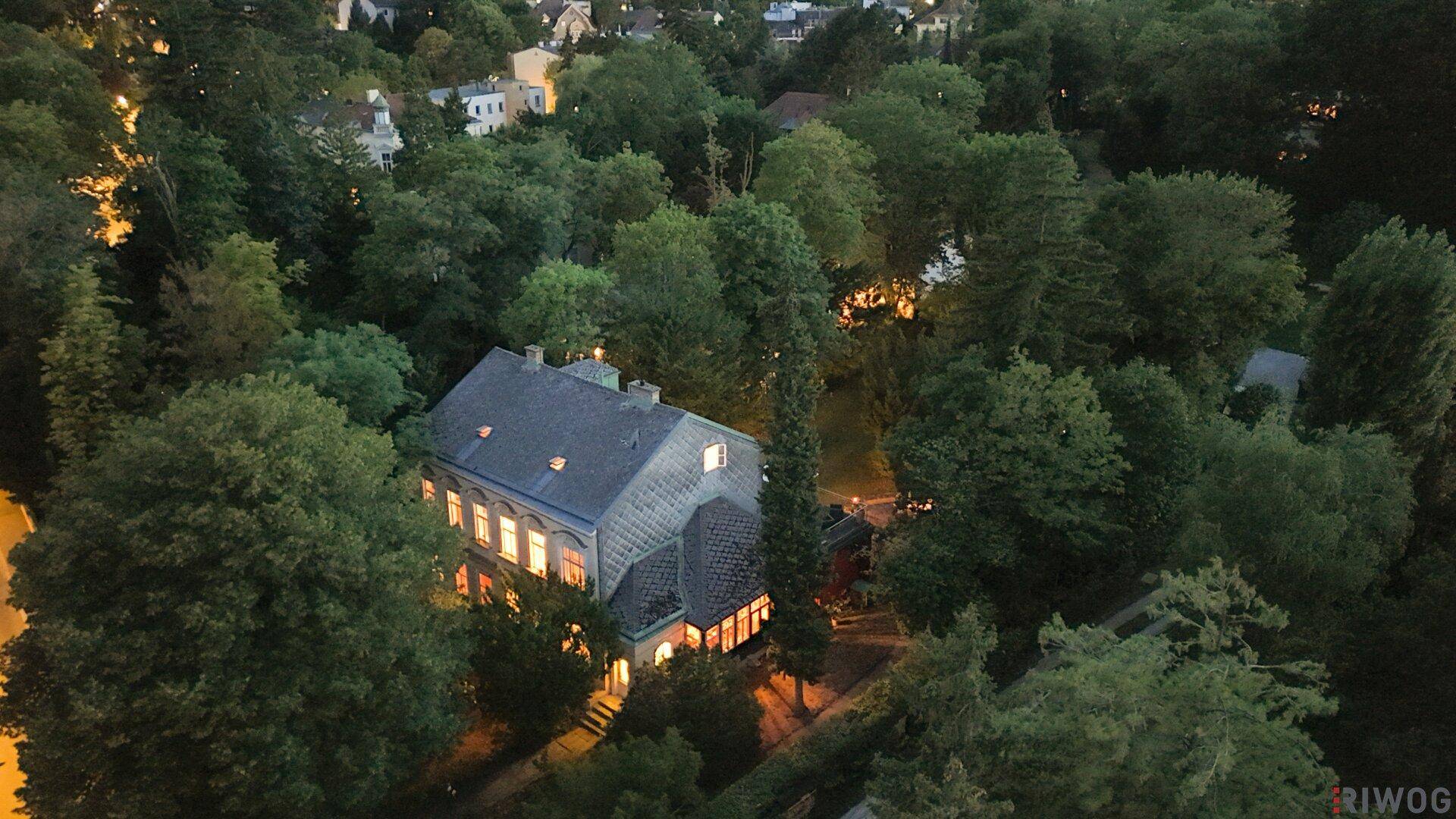 Luftaufnahme einer großen Villa, umgeben von dichtem Baumbestand, bei Dämmerung mit beleuchteten Innenräumen.