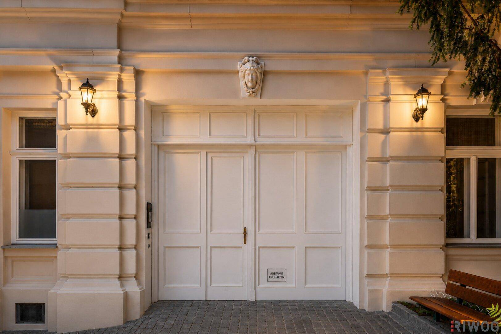 Elegante Gebäudefassade mit klassischem Eingangstor, flankiert von stilvollen Laternen und dekorativen Elementen.
