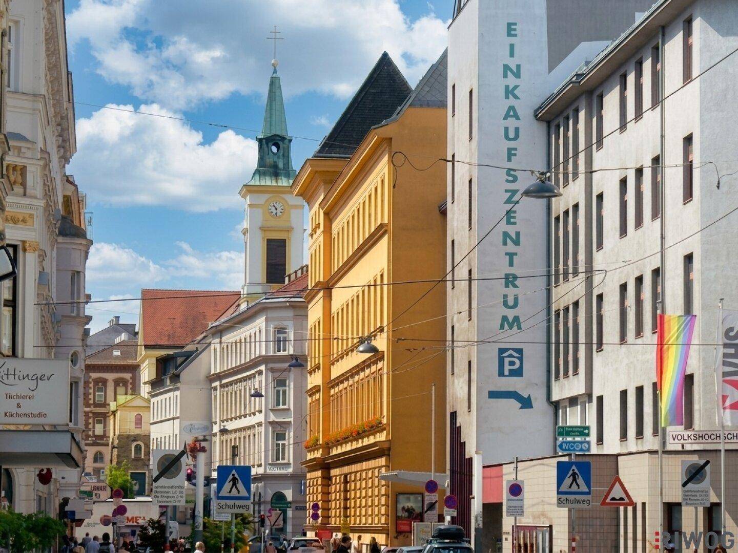 Belebte Stadtstraße mit historischen Gebäuden, einem Kirchturm und einem Einkaufszentrum unter blauem Himmel.