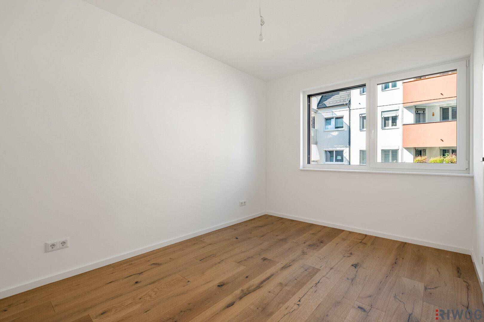 Leeres Zimmer mit Holzboden und einem großen Fenster, das viel Tageslicht hereinlässt.