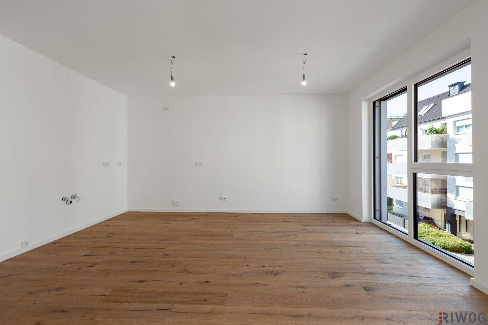 Helles, leeres Zimmer mit Holzboden und bodentiefen Fenstern, die viel Tageslicht hereinlassen.