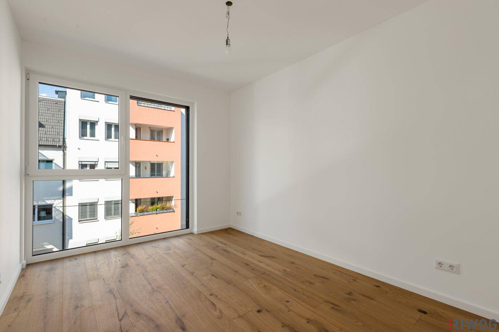 Leeres Schlafzimmer mit Holzboden und großem Fenster mit Blick auf die Nachbargebäude.