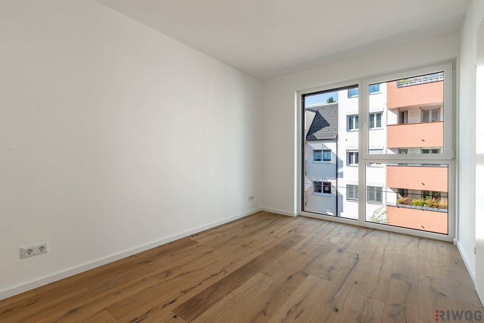Lichtdurchflutetes Zimmer mit Holzboden und großem Fenster, das einen Blick auf die Nachbarschaft bietet.