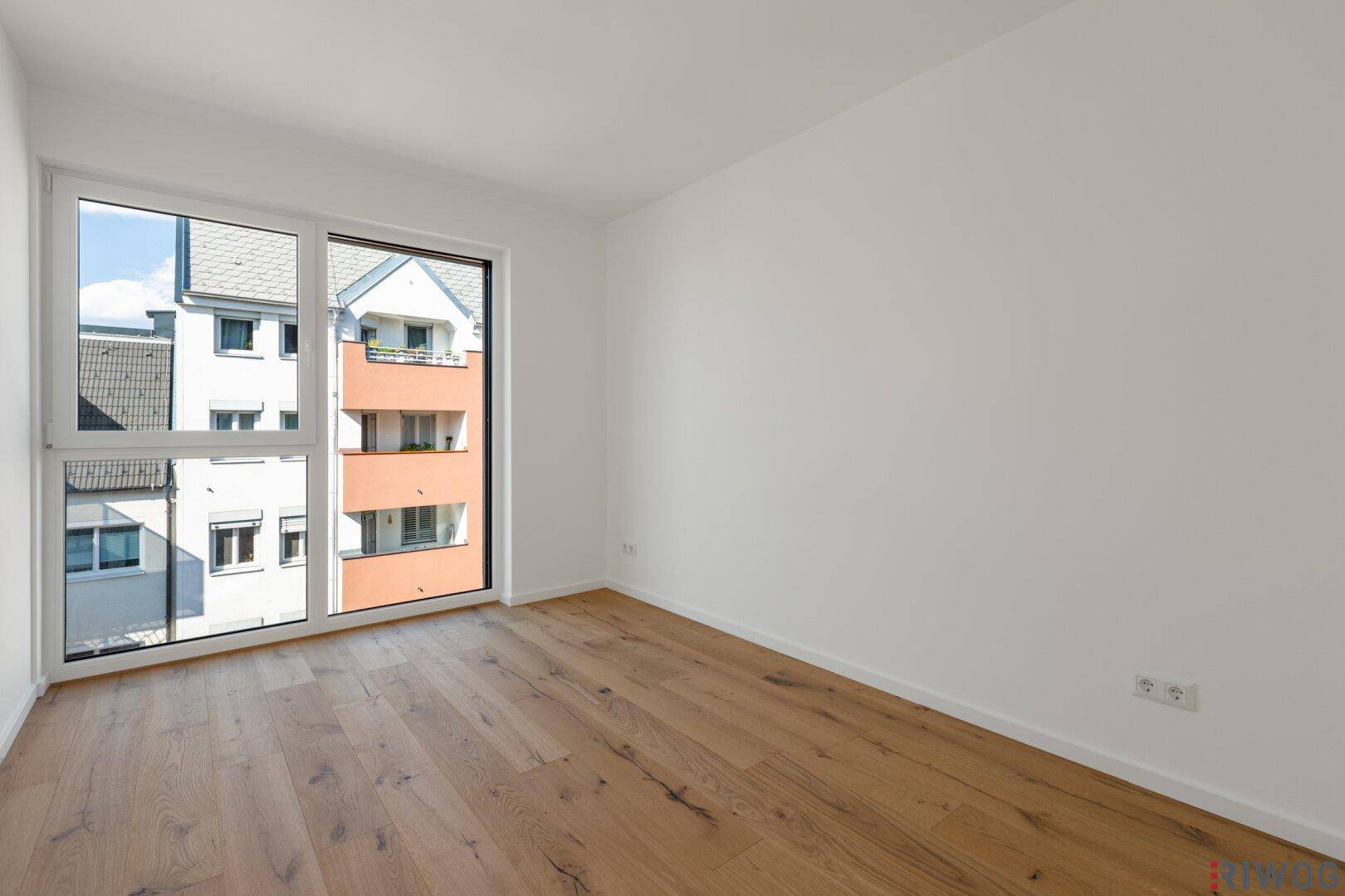 Helles Schlafzimmer mit Holzboden und großem Fenster mit Blick auf die Nachbargebäude.