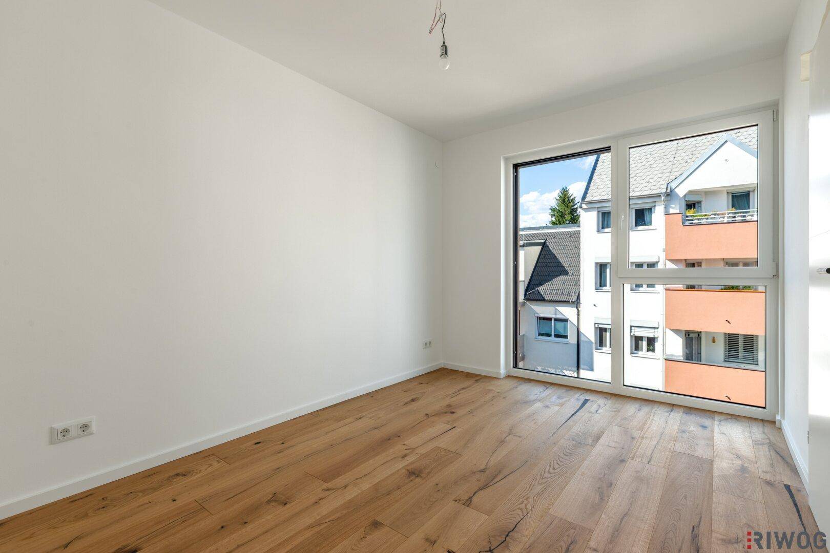 Helles, leeres Schlafzimmer mit Holzboden und großen Fenstern, bereit zur individuellen Einrichtung.