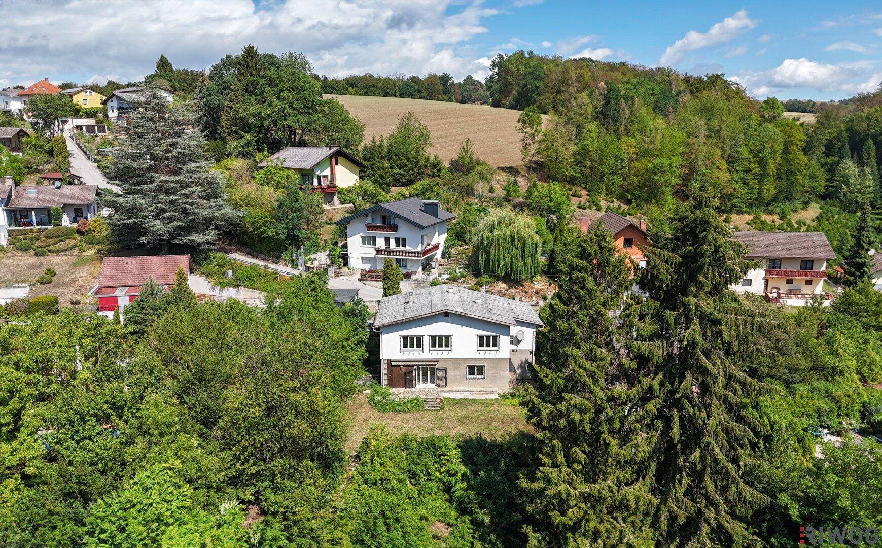 Luftaufnahme einer Wohnsiedlung in hügeliger Landschaft mit viel Grün und Wald.