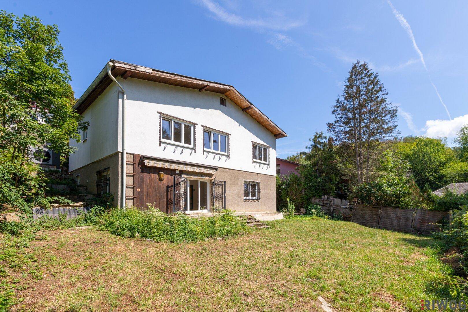Hausfassade mit großem, ungestaltetem Gartenbereich und viel Grün im Hintergrund.
