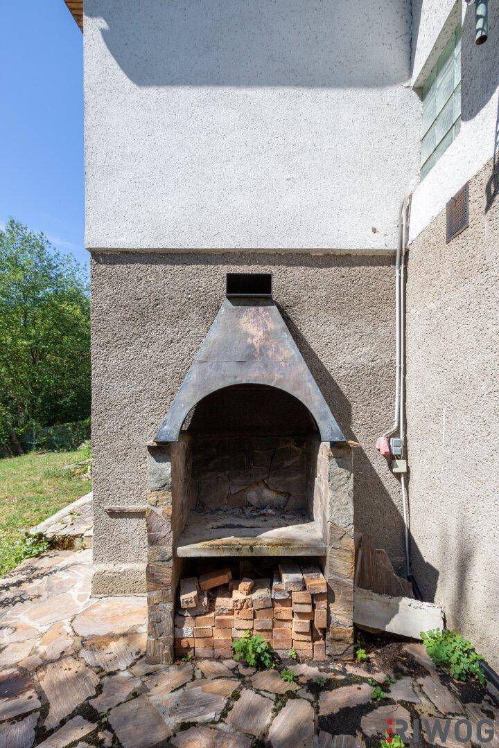 Gemauerter Außengrill mit Holzvorrat auf einer Steinfläche neben dem Haus.