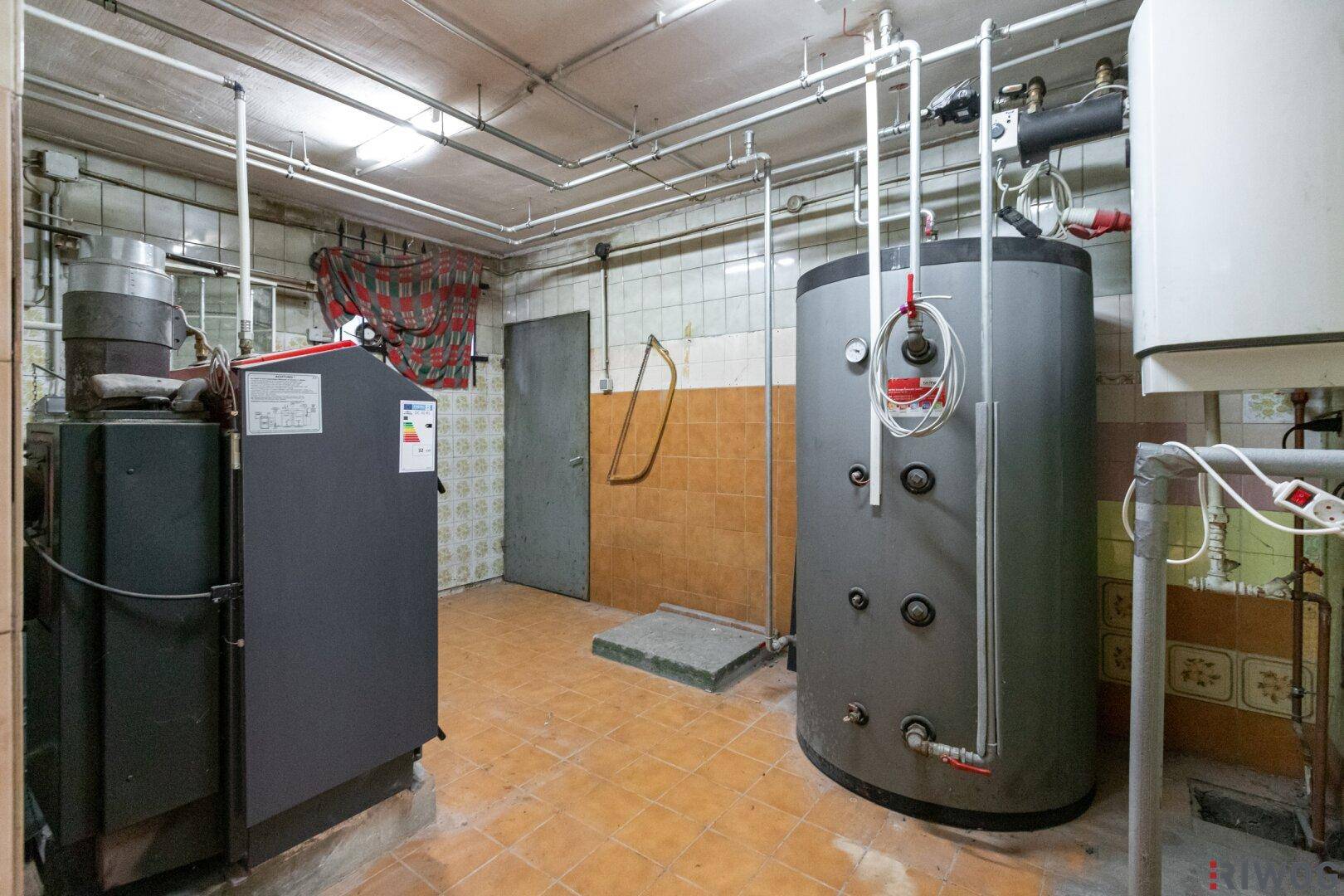 Heizungsraum im Keller mit großem Warmwasserspeicher und sichtbaren Rohrleitungen an gefliesten Wänden.