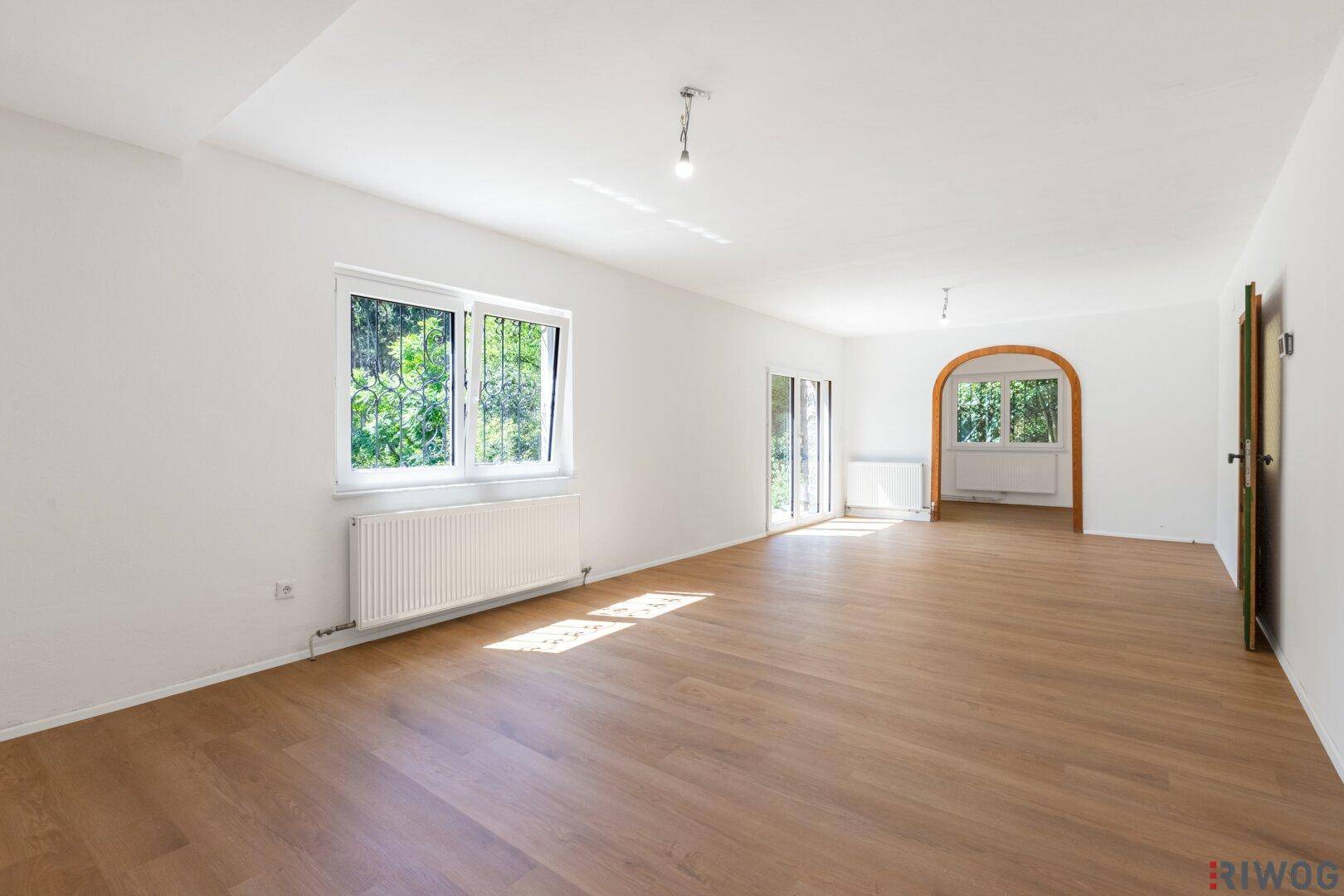 Helles, leeres Wohnzimmer mit Laminatboden und Blick ins Grüne.