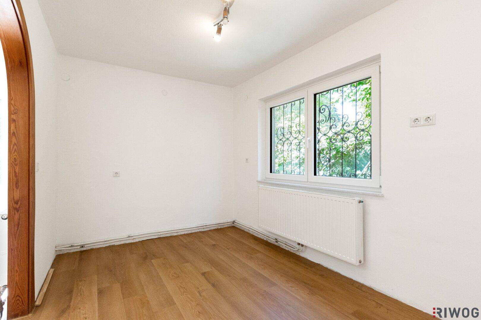 Helles, leeres Zimmer mit Laminatboden und Fenster zum grünen Außenbereich.