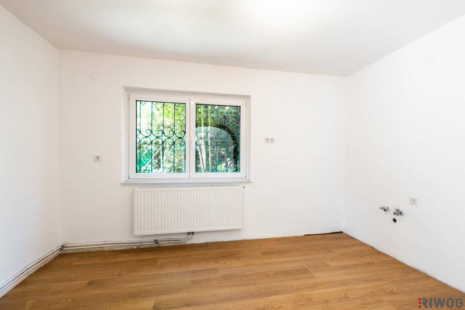 Leerer Raum mit Holzboden, weißen Wänden und Fenster mit Blick ins Grüne.