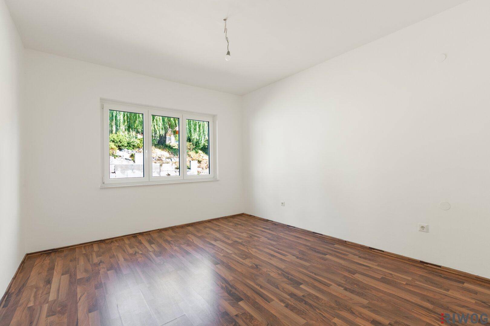 Leerer Raum mit Holzboden, weißen Wänden und Fenster mit Blick ins Grüne.