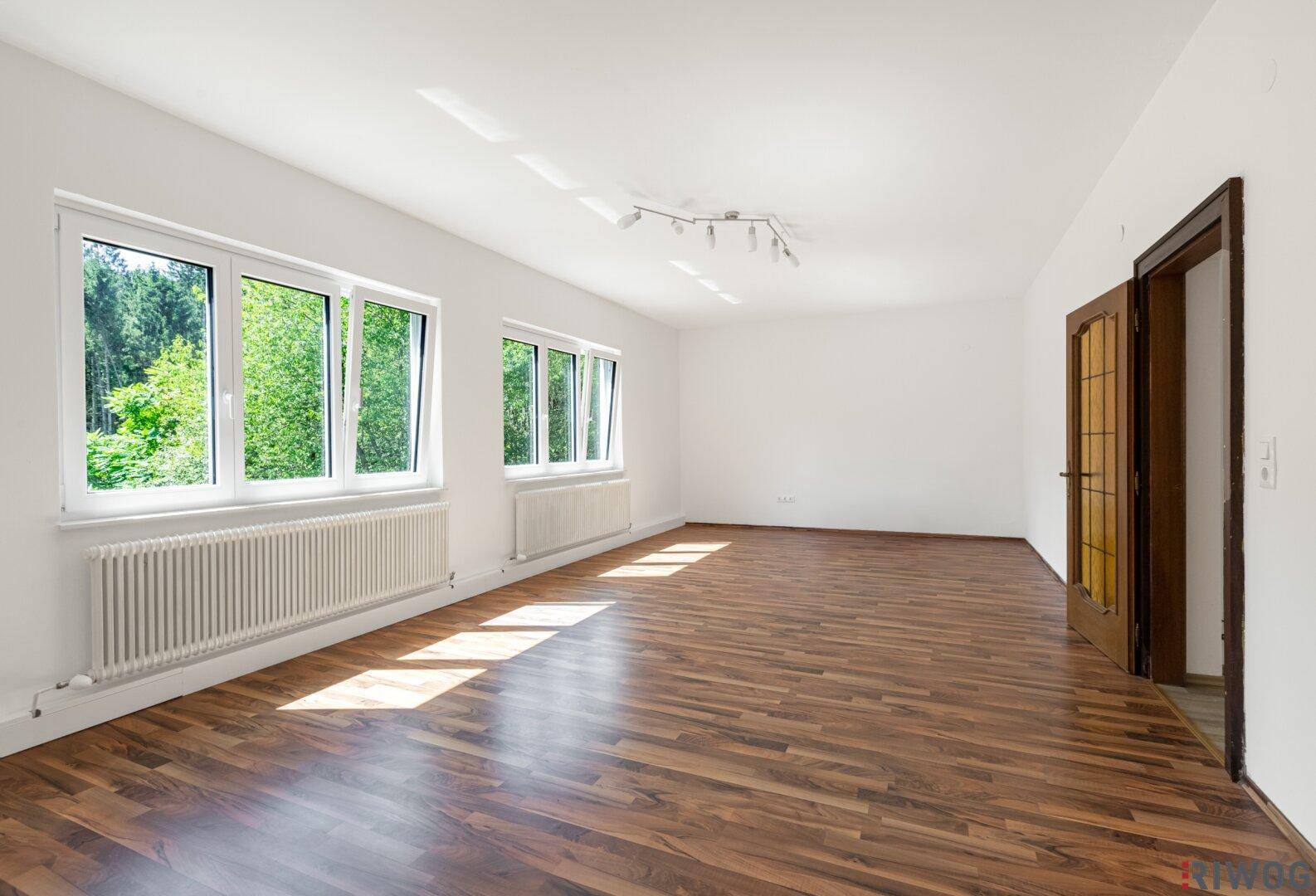 Großer, heller Raum mit Holzboden, weißen Wänden und Fenstern mit Waldblick.