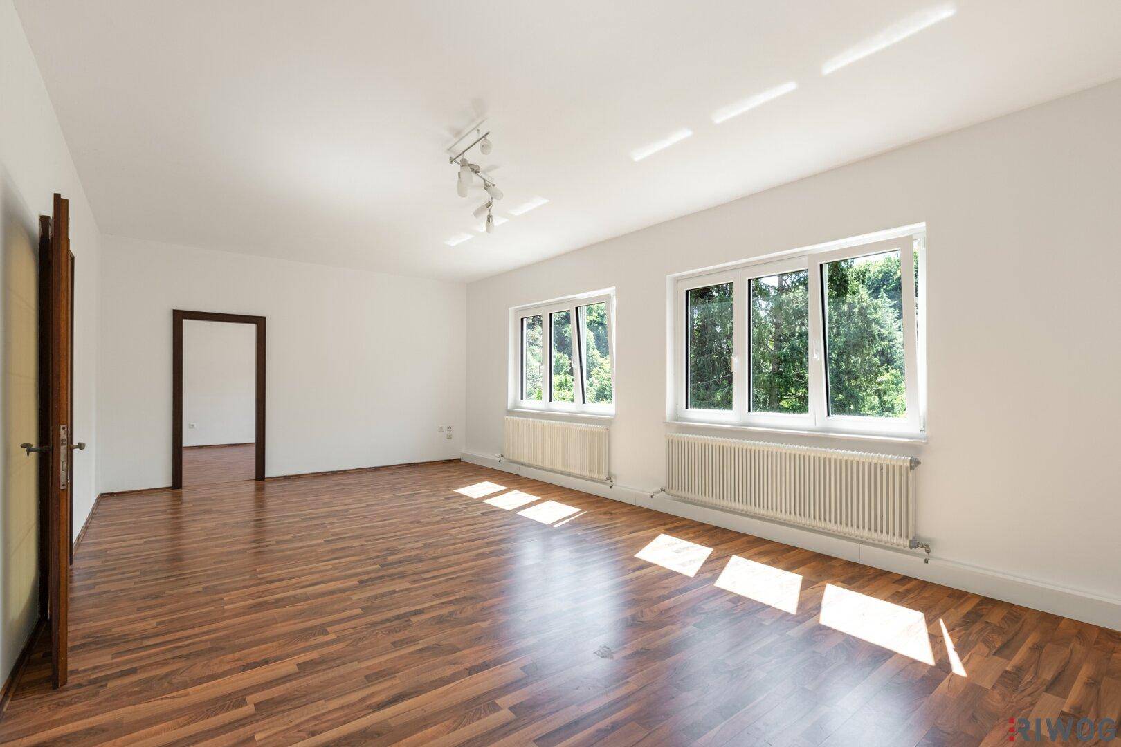 Heller Raum mit Holzboden, weißen Wänden und Fenstern mit Blick auf den Wald.