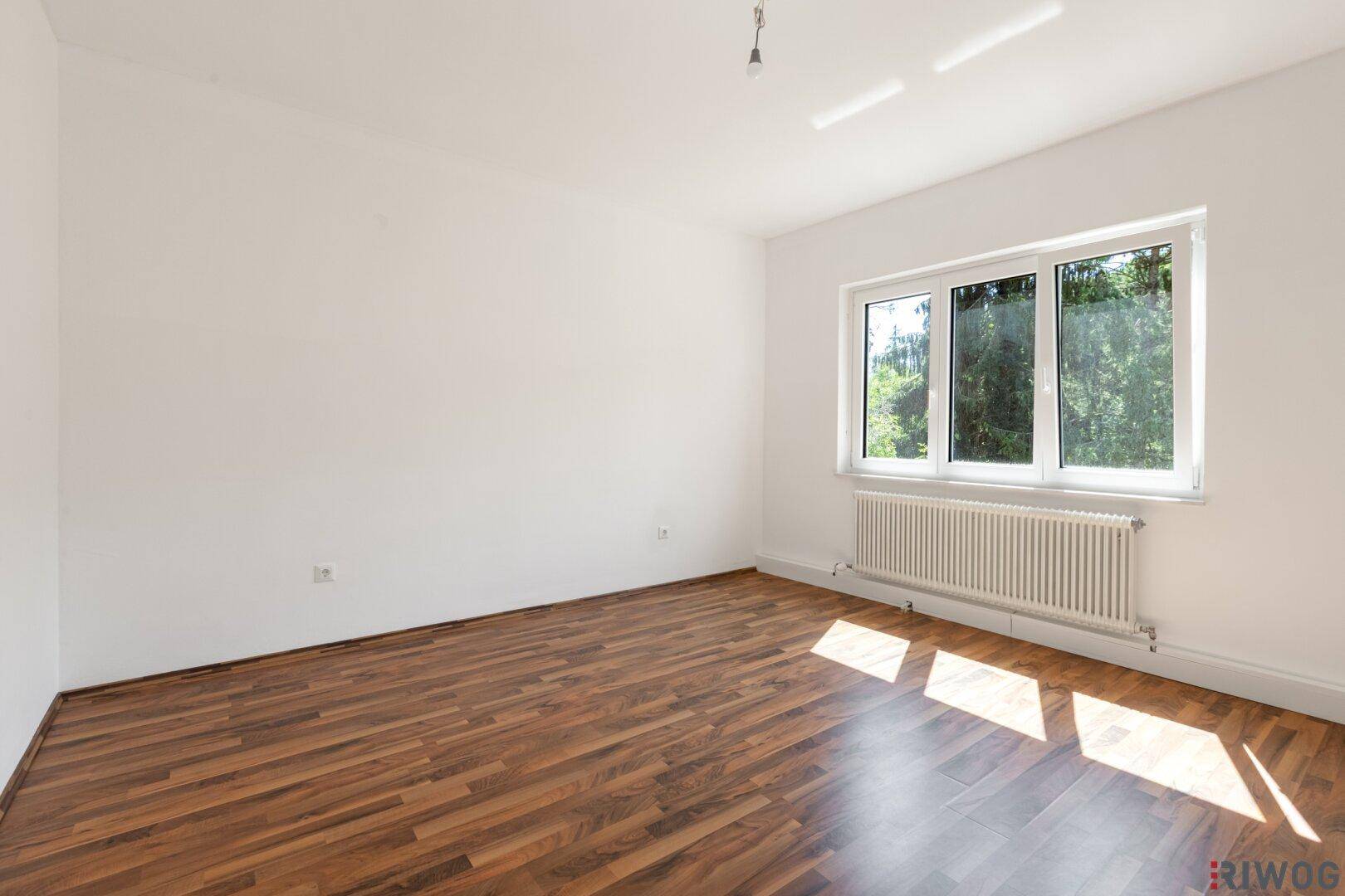 Leerer Raum mit Holzboden, weißen Wänden und Fenster mit Blick ins Grüne.