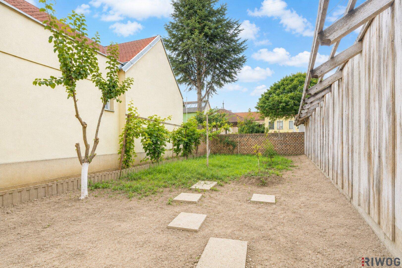 Gartenbereich mit jungen Bäumen, Trittsteinen und einer Ziegelmauer im Hintergrund.