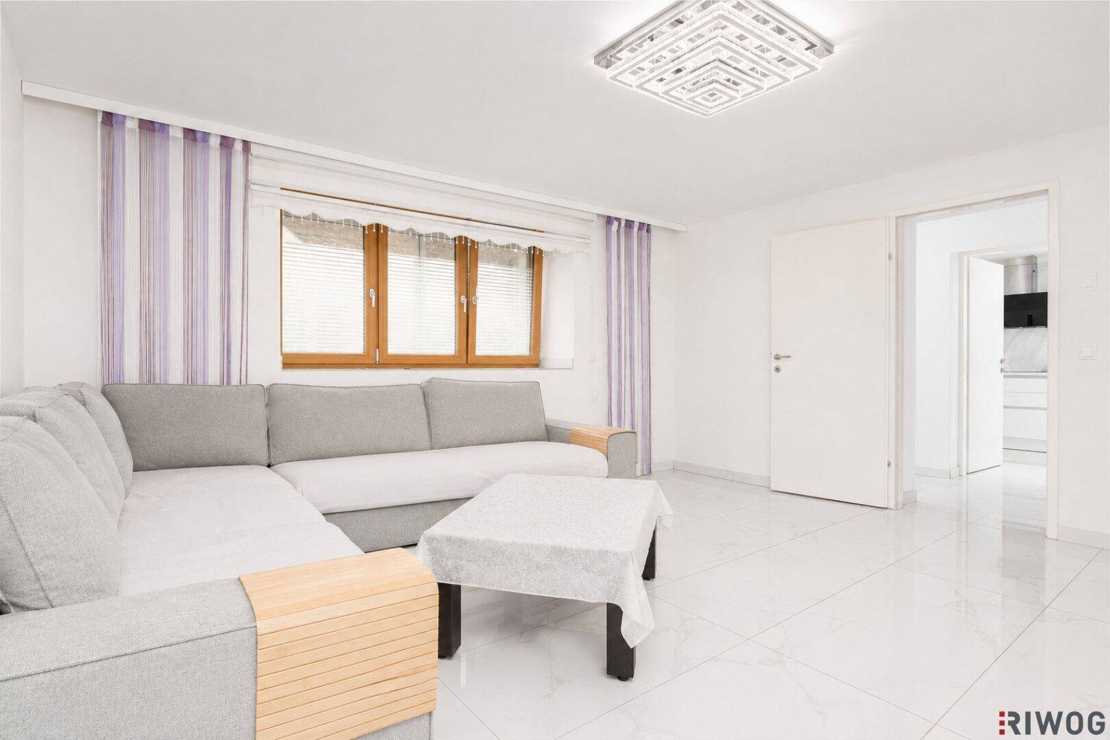 Helles Wohnzimmer mit großem Ecksofa, Fliesenboden und Blick in die moderne Küche.