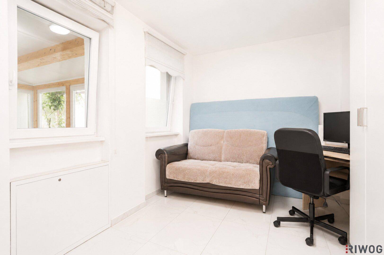 Helles Zimmer mit Sofa und Schreibtisch, mit Fenster zum Wintergarten.