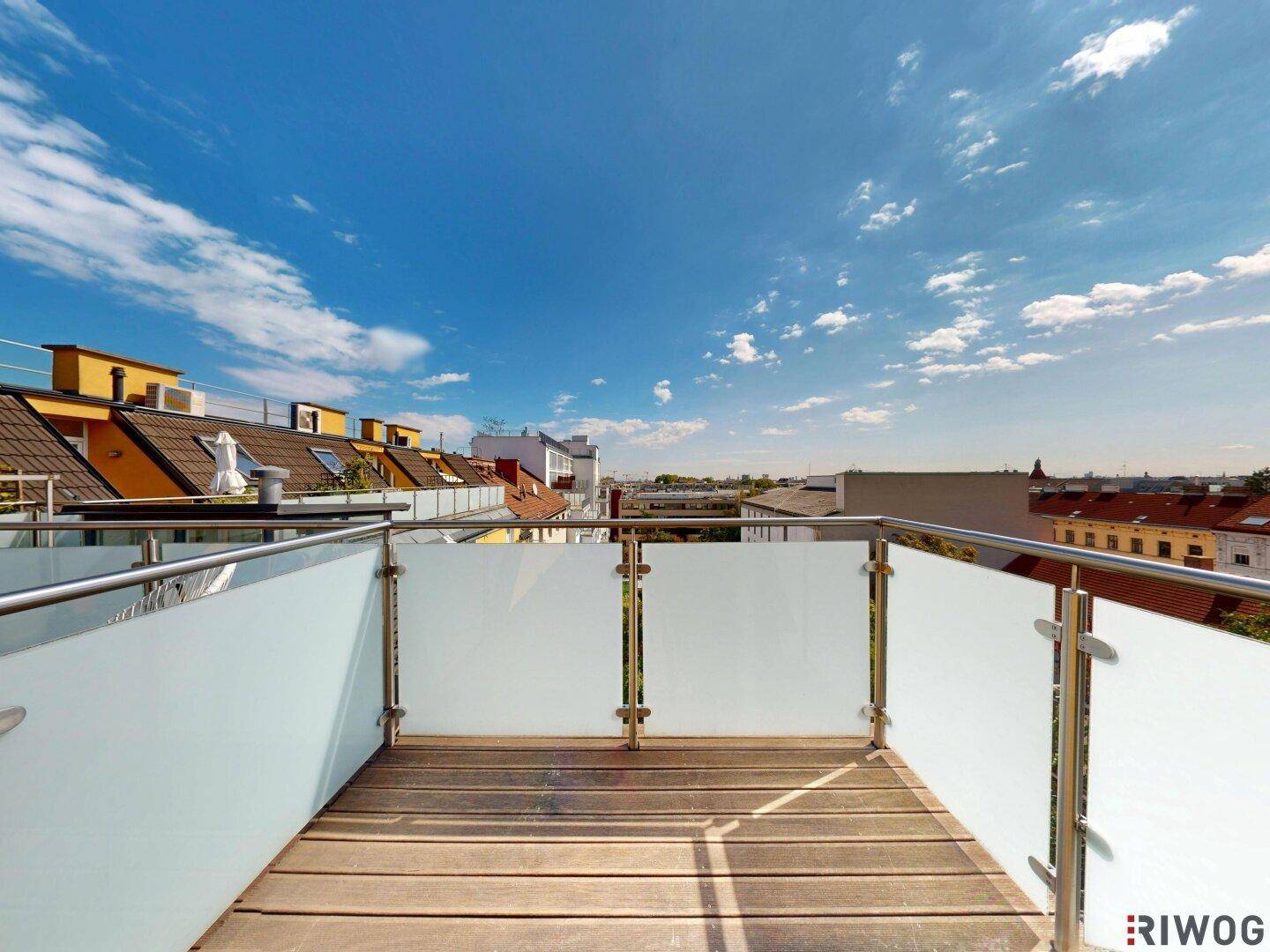 Heller Balkon mit Holzbelag und modernem Glasgeländer unter blauem Himmel.