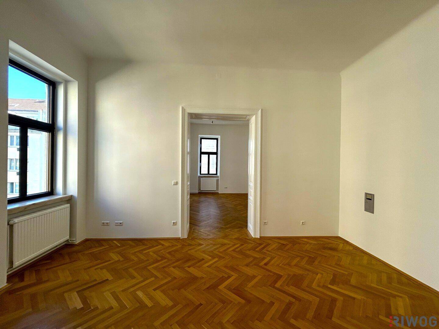 Lichtdurchflutetes Zimmer mit Fischgrätparkett, hohen Decken und Blick in einen weiteren Raum.