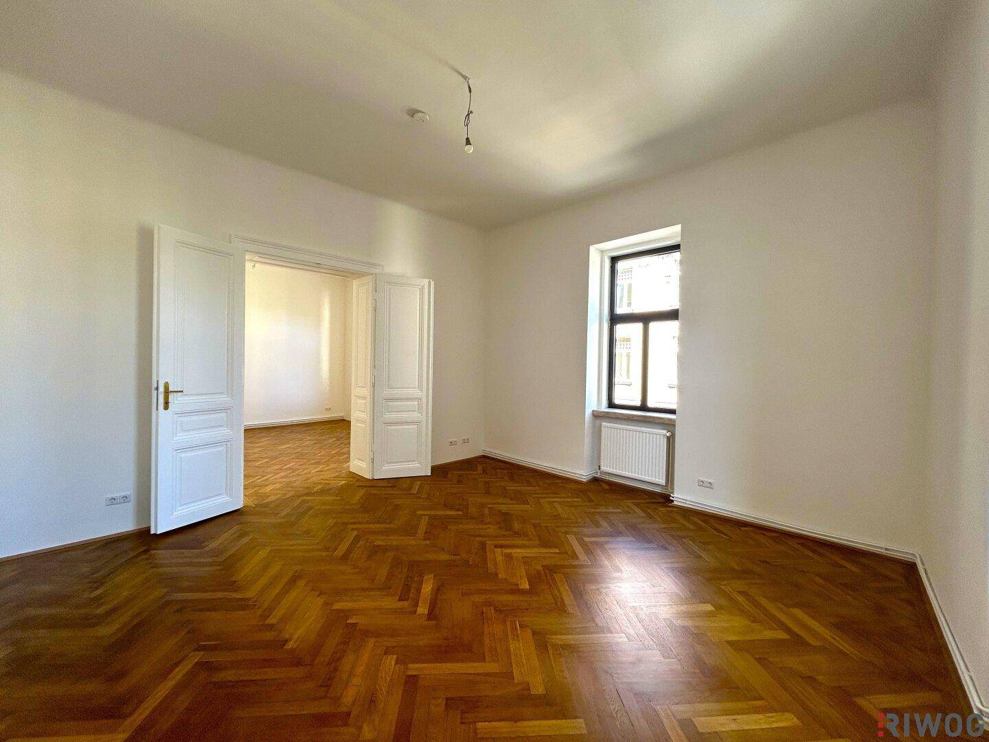 Helles Zimmer mit Fischgrätparkett, weißen Wänden und klassischen Doppeltüren, die den Raum öffnen.