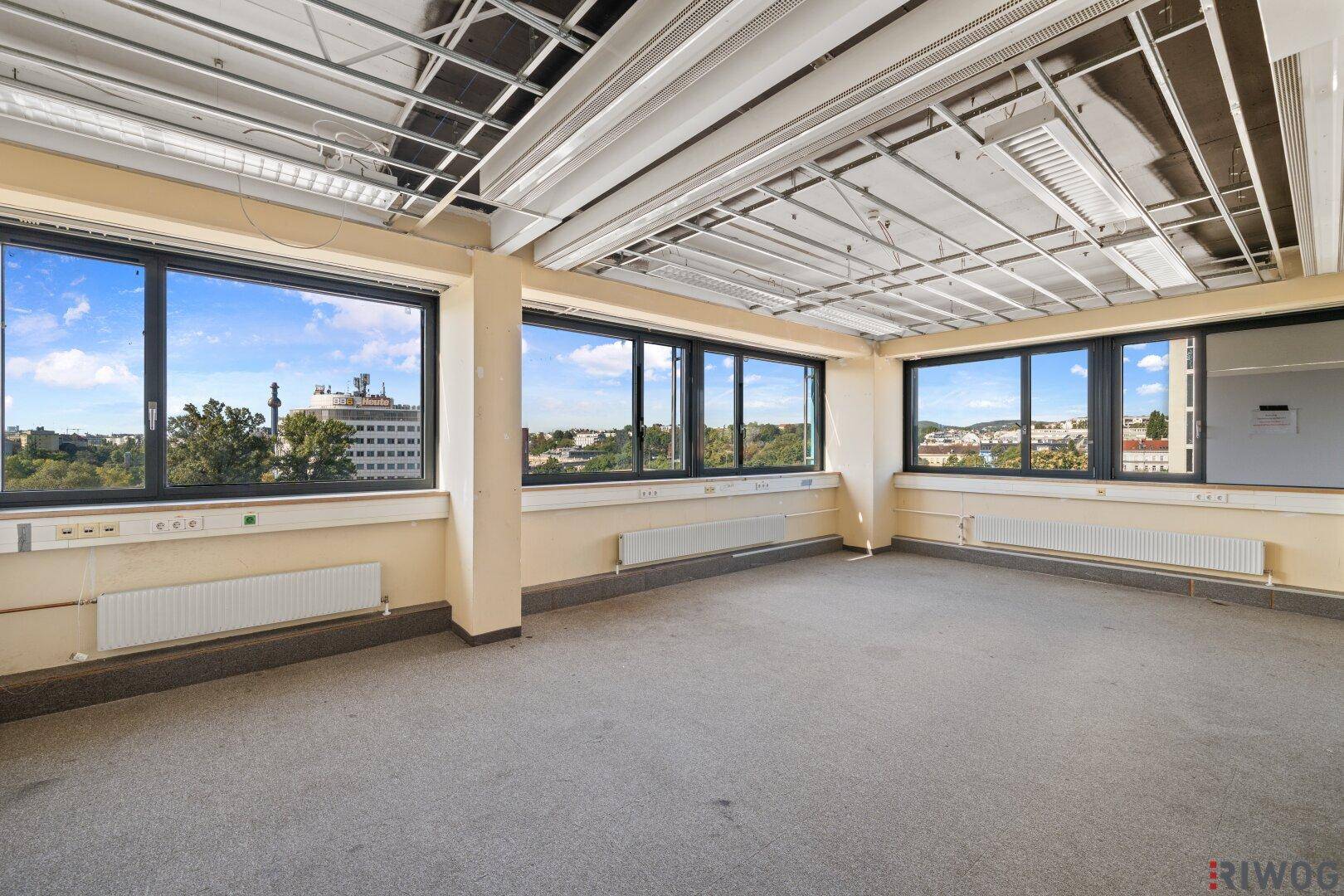 Leerer Büroraum mit Teppichboden, großen Fenstern und Blick auf die Stadt.