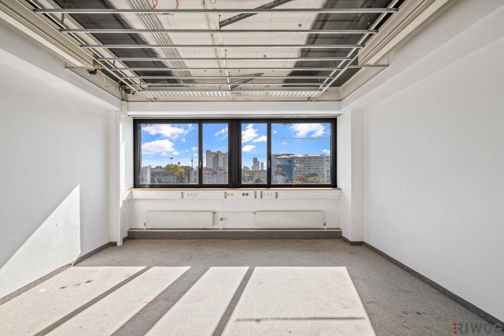 Leerer Büroraum mit Teppichboden, großen Fenstern und Blick auf die städtische Skyline.