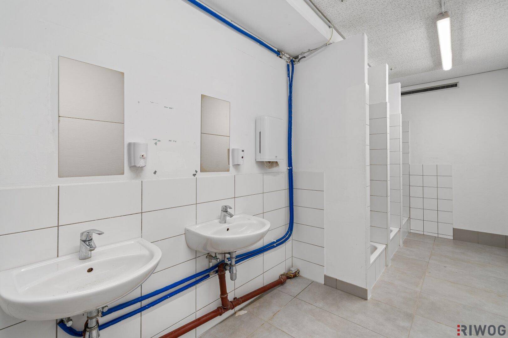 Sanitärbereich mit mehreren Waschbecken, Fliesen und sichtbaren Wasserleitungen.