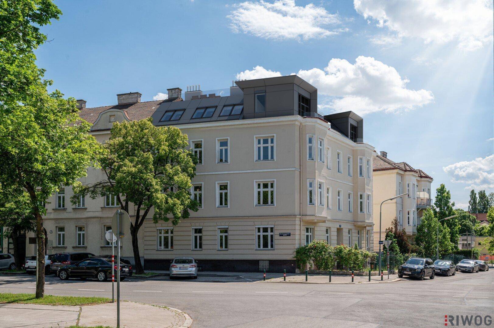 Ansicht eines renovierten Eckgebäudes mit modernem Dachausbau und grüner Umgebung an einem sonnigen Tag.