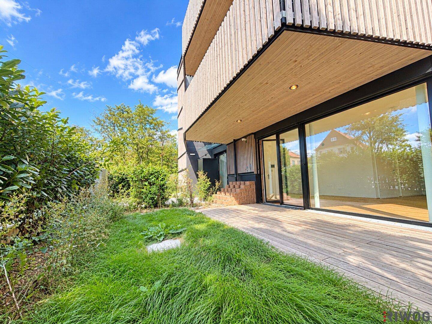 Die moderne Holzterrasse grenzt an das Gebäude mit großen Glasfronten und umgebendem Grün.