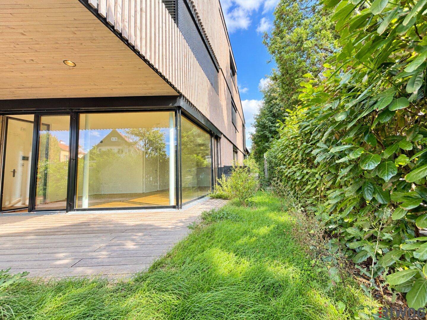 Seitlicher Blick auf die Holzterrasse und die moderne Fassade, umgeben von üppigem Grün.
