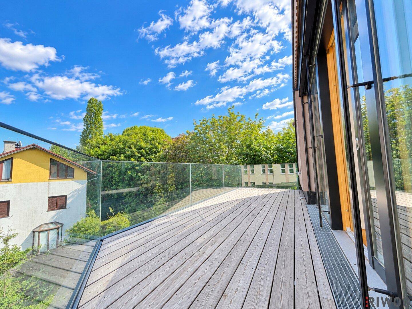 Geräumige Holzterrasse mit Glasgeländer und Blick auf die umliegende Bebauung und viel Grün.
