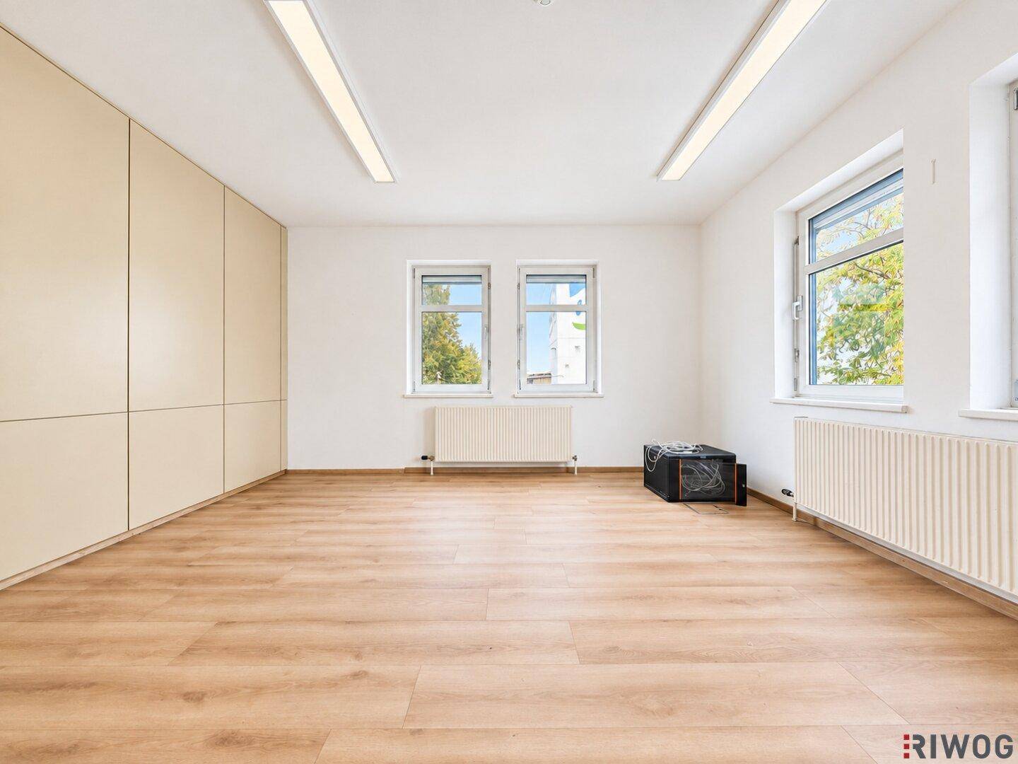Leeres Büro mit großen Fenstern, Heizkörpern und einem hellen Laminatboden, bereit zur Einrichtung.