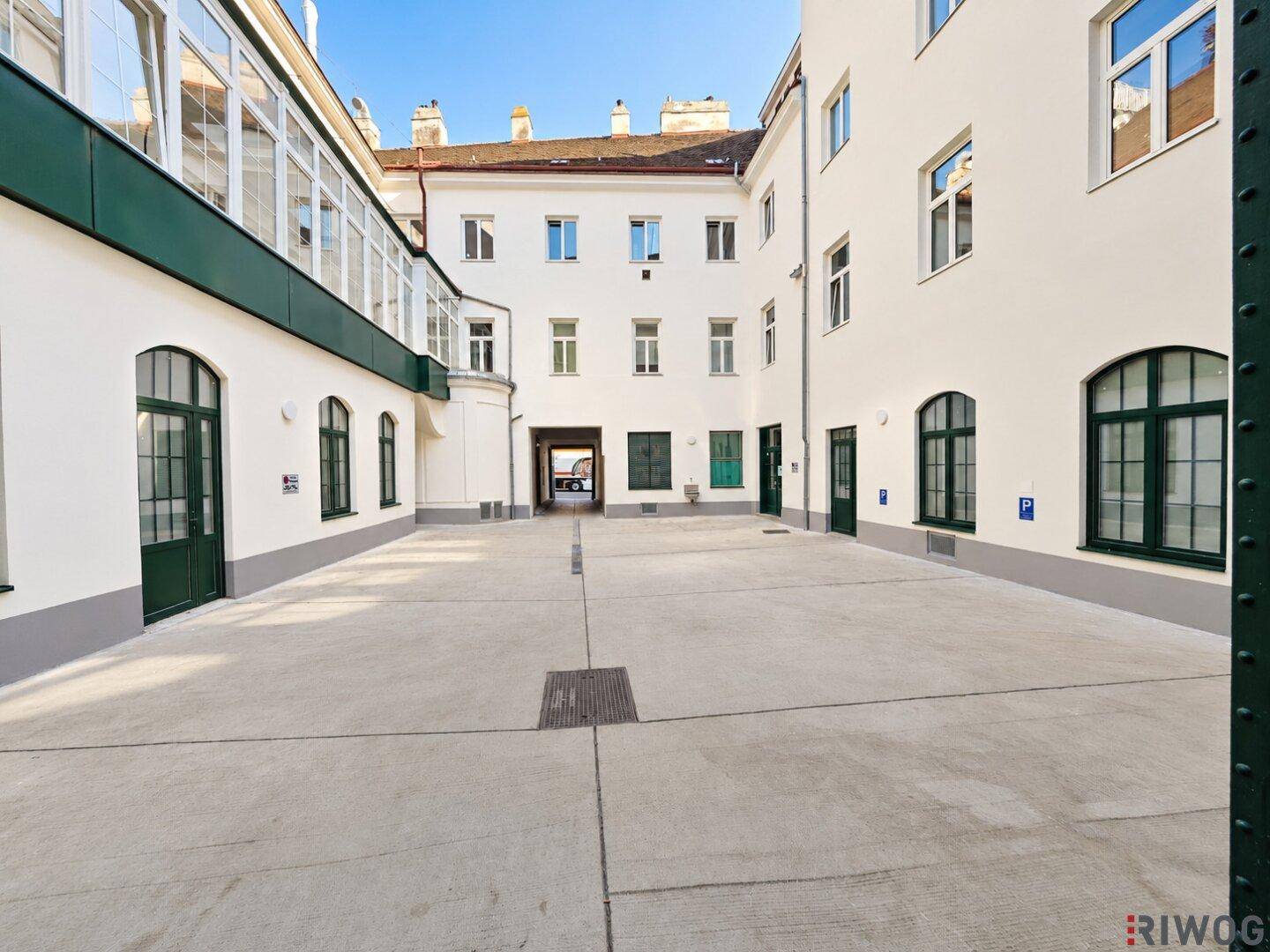 Heller Innenhof mit Betonboden, umgeben von Gebäuden mit weißen Fassaden und grünen Fensterrahmen.