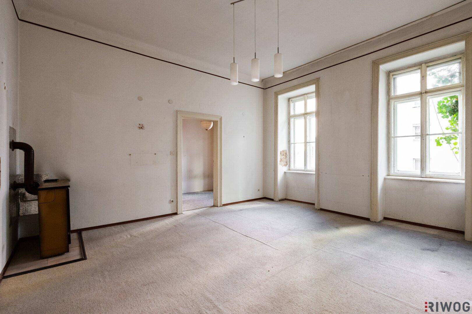 Leeres Zimmer mit historischem Ofen, zwei Fenstern und Teppichboden, in sanierungsbedürftigem Zustand.