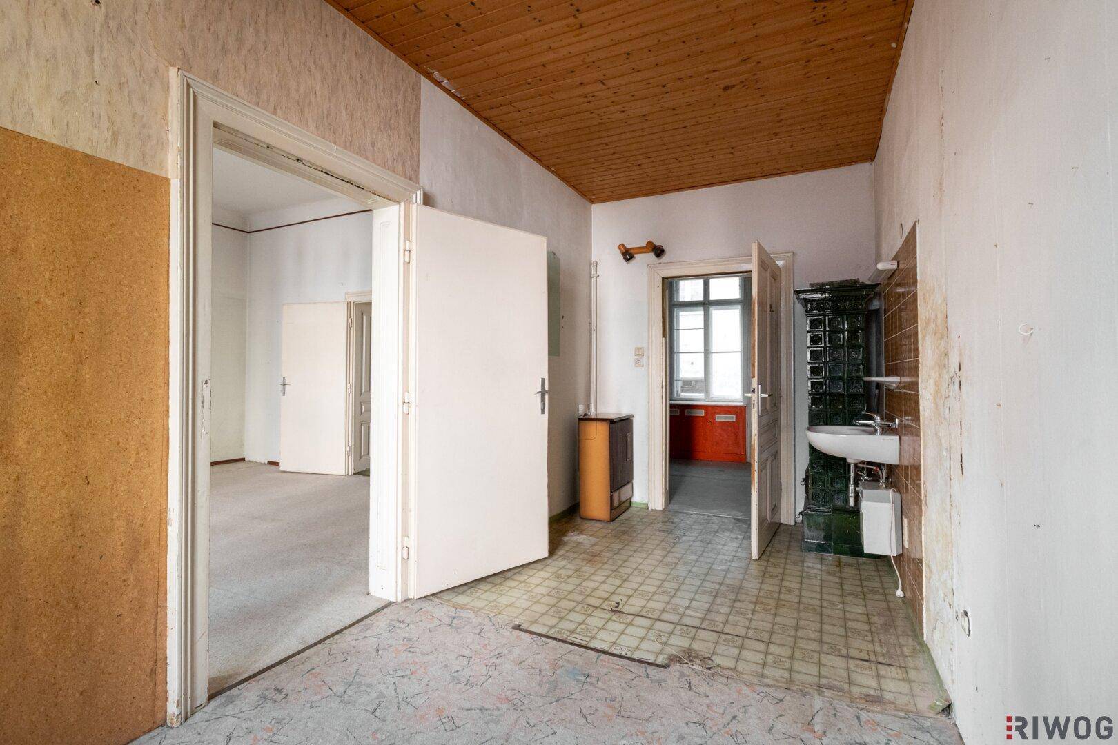 Flur mit Waschbecken und Kachelofen, der zu weiteren Räumen führt, in sanierungsbedürftigem Zustand.