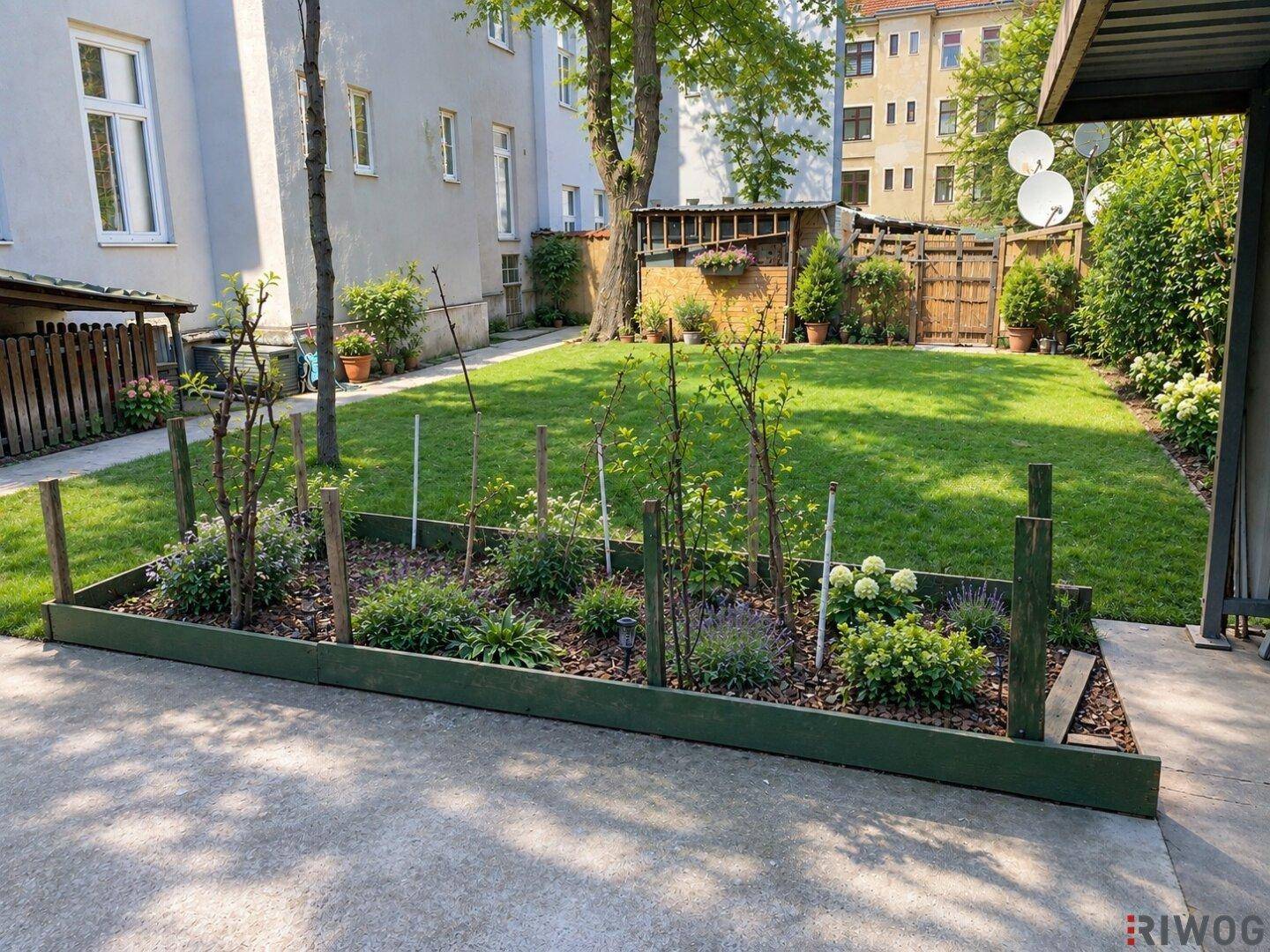 Gepflegter Gemeinschaftsgarten mit Rasenfläche, Hochbeet und altem Baumbestand im Hintergrund.