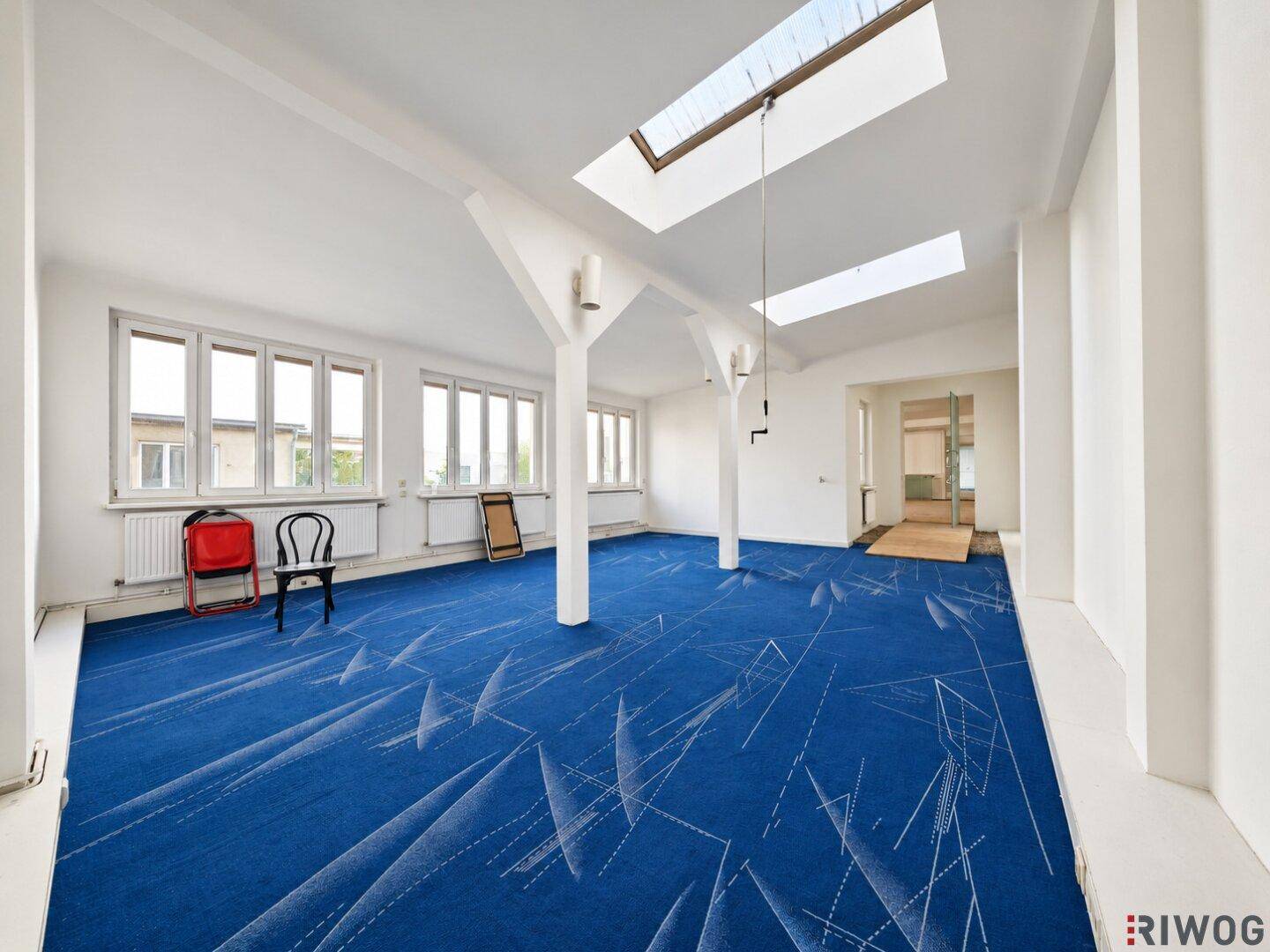 Heller Büroraum mit auffälligem blauem Teppich, Oberlichtern und großen Fenstern für viel Tageslicht.