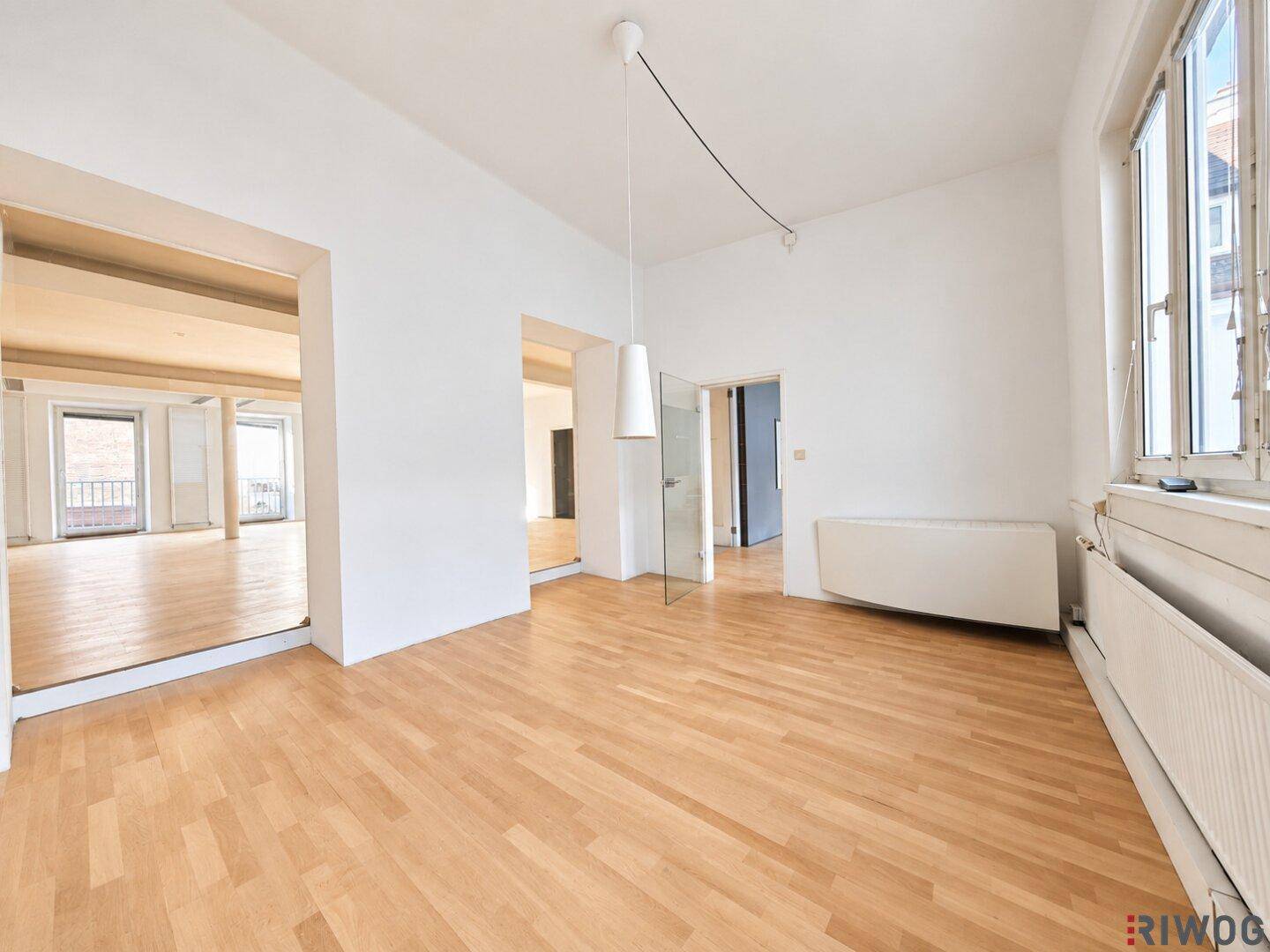 Leerer Raum mit Holzboden und breitem Durchgang zu einem weiteren hellen Bereich mit Fenstern.