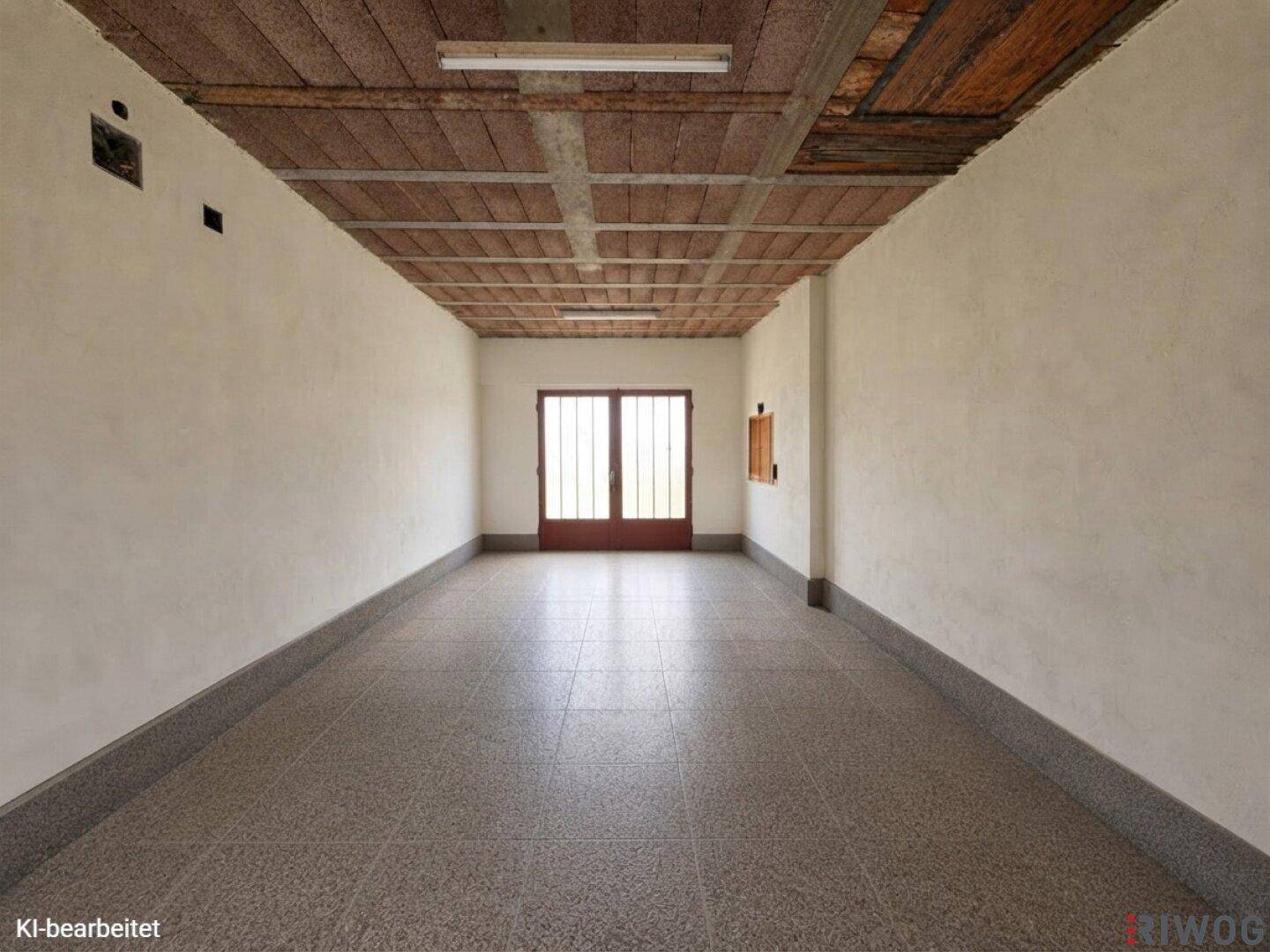 Langer, leerer Raum mit Fliesenboden, Holzbalkendecke und Doppeltüren am Ende.
